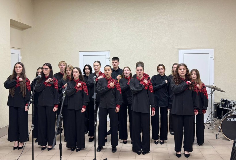 Хористи у чорно-червоних костюмах співають та тримають руку на серці