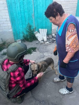 Дві жінки, одна з них волонтер у захисній екіпіровці, стоять біля пса, якого звуть Мухтар, волонтер тримає його за ошийник.