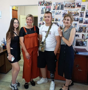 Група з чотирьох людей позує і посміхається в кімнаті зі стіною, обклеєною фотографіями. Настрій веселий і дружній. Серед них є молодий чоловік з протезом ноги.