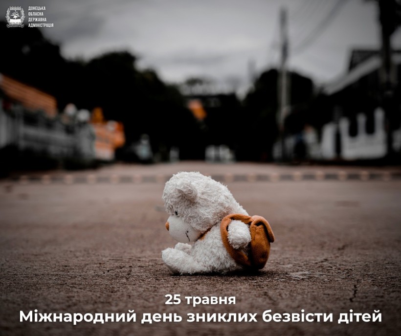 На вулиці покинутий ведмедик сидить на асфальті. На тлі видно міські будівлі та дерев'яні огорожі.