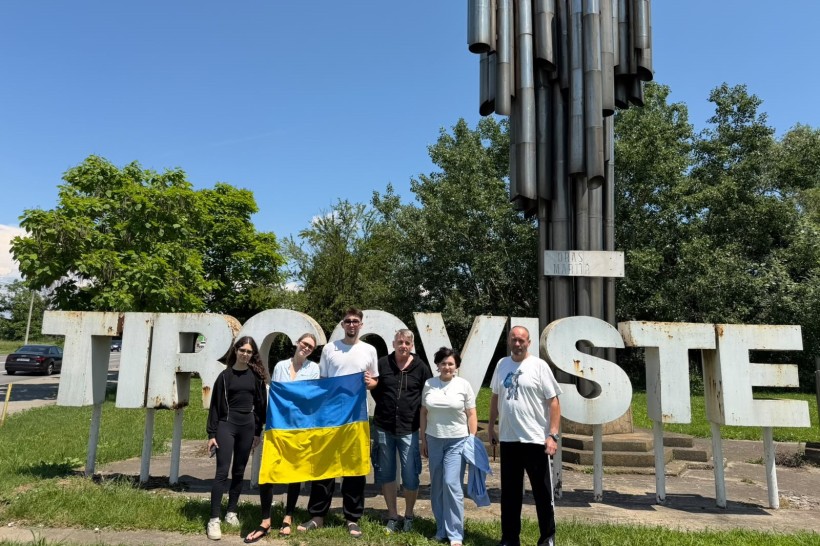 Театральна трупа позирує на тлі великого білого напису "Targoviste" та металевої стели, тримаючи прапор України