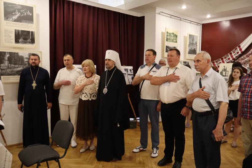 Відвідувачі на виставці, серед яких священнослужителі, тримають руку на серці під час офіційної частини заходу