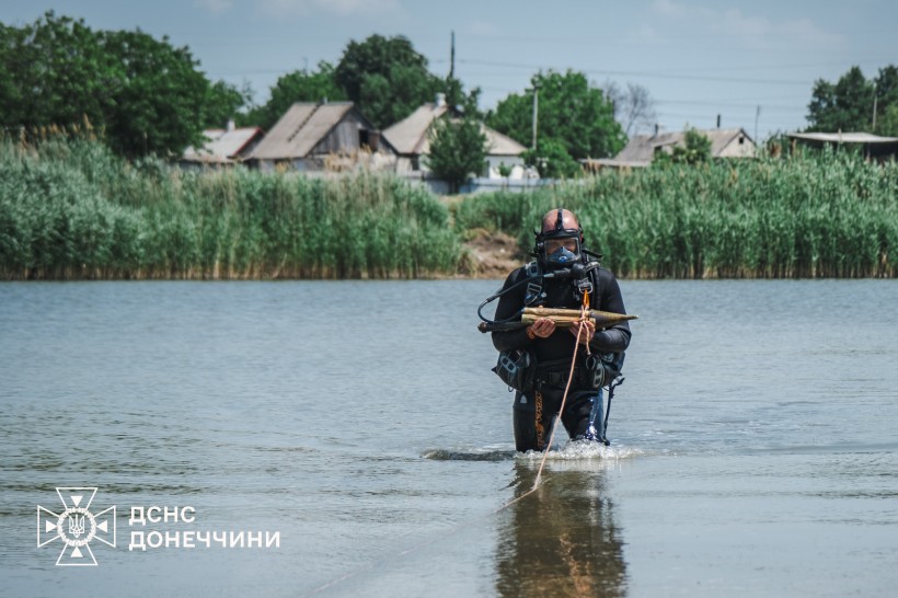 Розмінувальник служби ДСНС несе обережно з водойми артилерійський снаряд. Позаду водойми знаходяться житлові будинки.