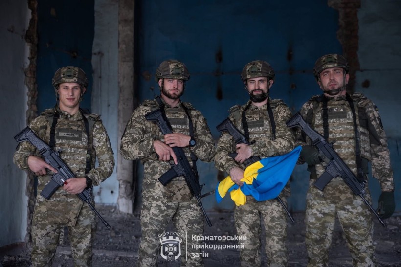 Четверо солдатів у камуфляжі та шоломах тримають гвинтівки в слабо освітленій кімнаті. Один з них тримає синьо-жовтий прапор. Вони стоять впевнено, випромінюючи рішучість.