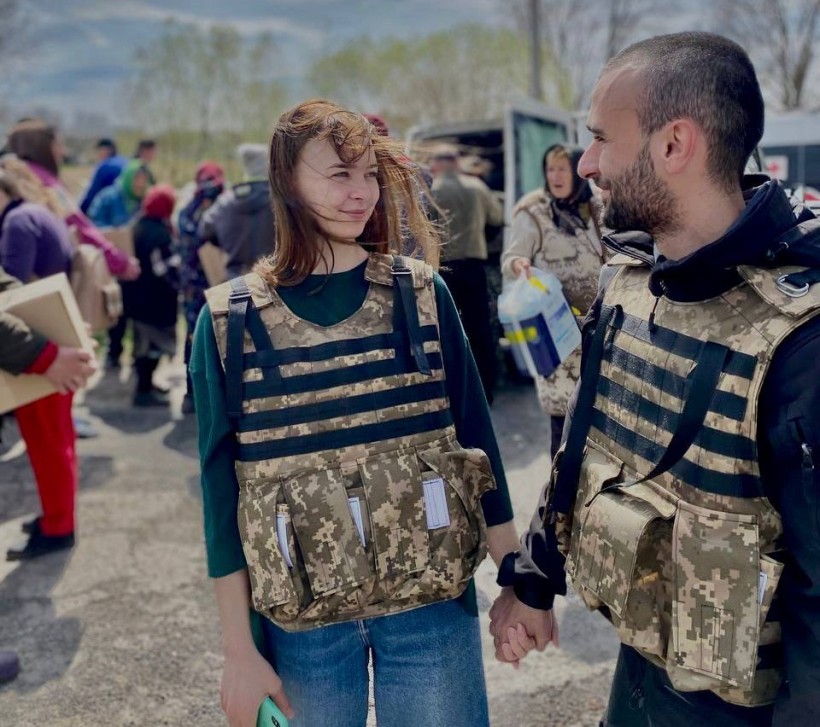 Молоді жінка та чоловік у бронежилетах спілкуються на фоні людей, які отримують гуманітарну допомогу