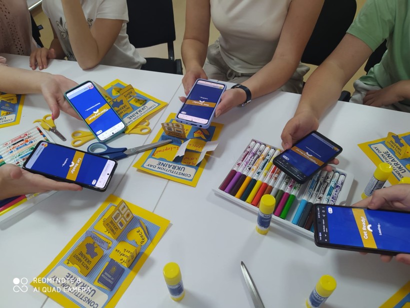 Учасники заходу сканують QR-код для онлайн-вікторини до Дня Конституції України. На столі — тематичні лепбуки та канцелярія.