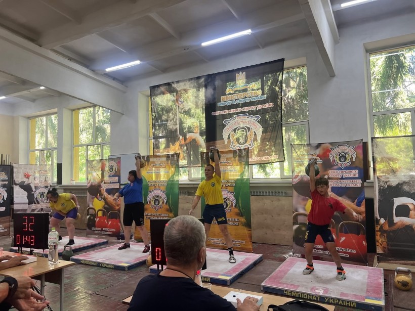 Спортсмени з гирьового спорту. В руках у спортсменів гирі, які вони підіймають однією рукою над головою. Учасники одягнені у спортивний одяг.