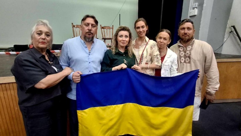 Актори та організатори вистави спільно тримають великий прапор України на сцені біля стільців.