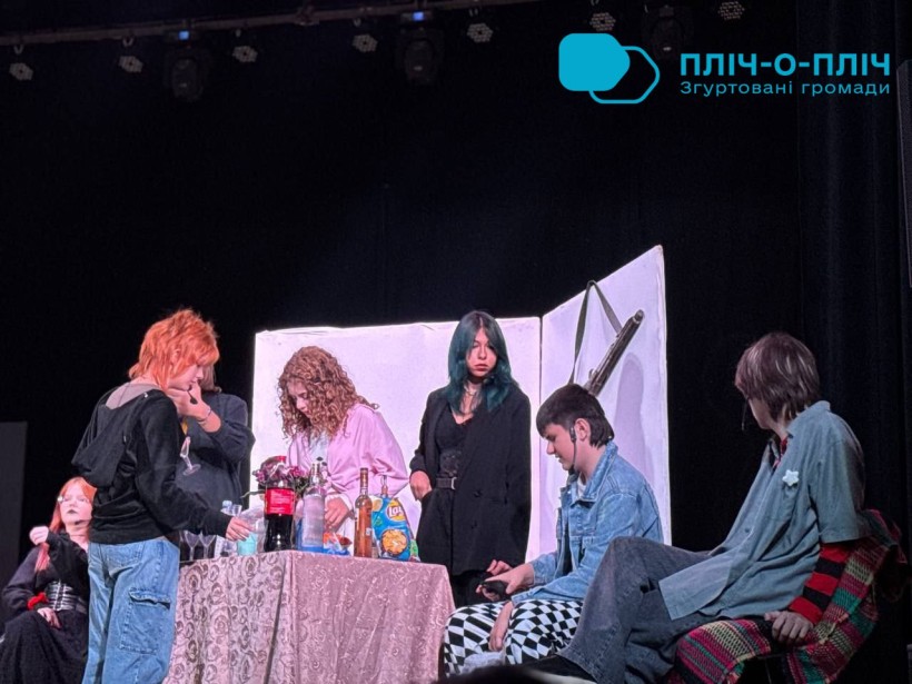A group of five people gather around a table on stage, focused on various activities. The setting has a relaxed, theatrical atmosphere with soft lighting.