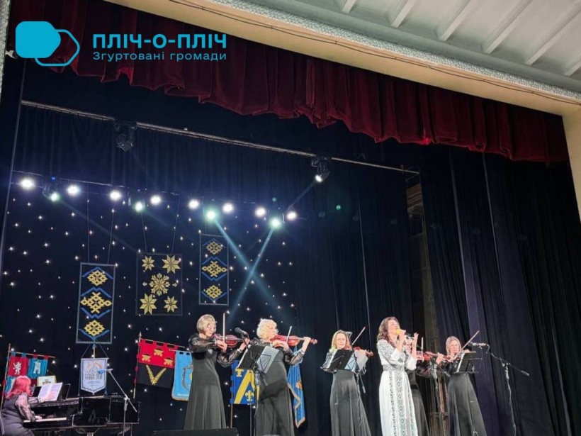 Дія на сцені. Жінки у чорних сукнях тримають скрипки та грають. Жінка у сірому платті співає у мікрофон.