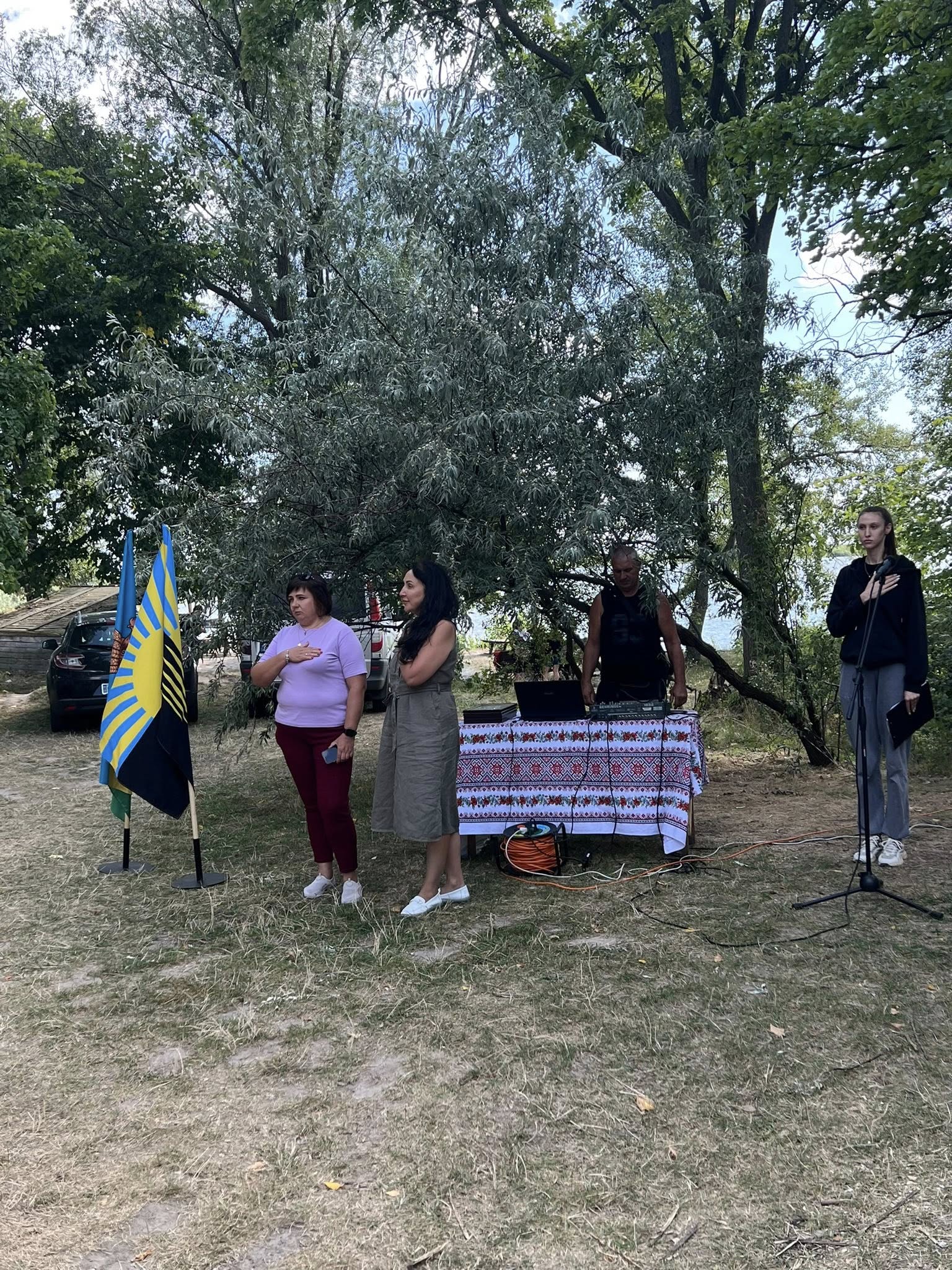 Заступник начальника Шахівської сільської військової адміністрації Лілія Тихонова та інші учасники співають гімн України. Руки біля серця. Поруч з нею прапори Донеччини та громади.