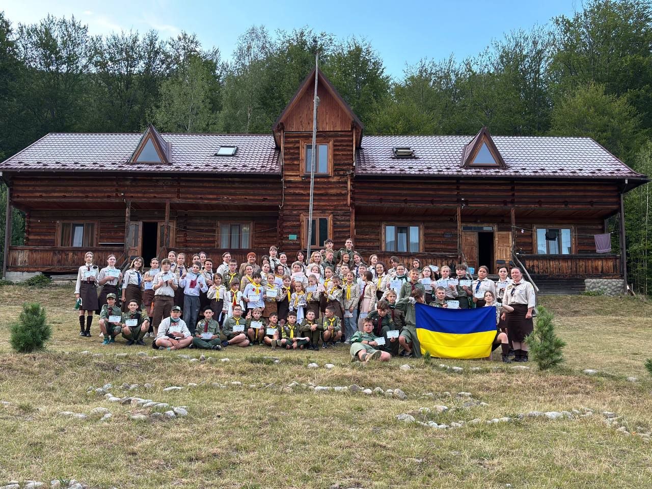 Учасники пластового табору «Земля велетнів» тримають український прапор перед дерев'яним будинком.