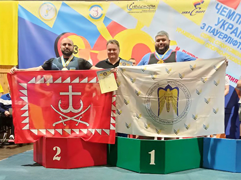 Спортсмени пара пауерліфтери, тримають прапори по центру стоїть тренер.