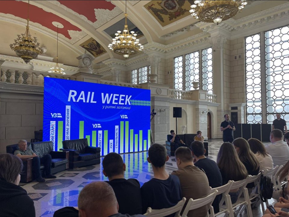 Аудиторія слухає виступ спікера на ярмарку вакансій RAIL WEEK - у ритмі залізниці' у великій залі з високою стелею та люстрами. На величезному синьому екрані позаду спікера відображається назва заходу та графіки.
