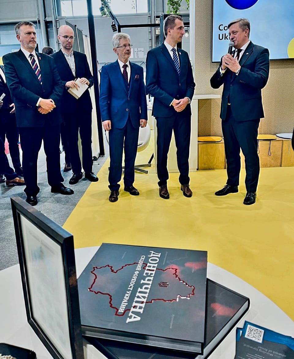 Учасники форуму. Чоловіки у ділових костюмах, один з них тримає мікрофон та говорить. На столі лежать книги про Донеччину.