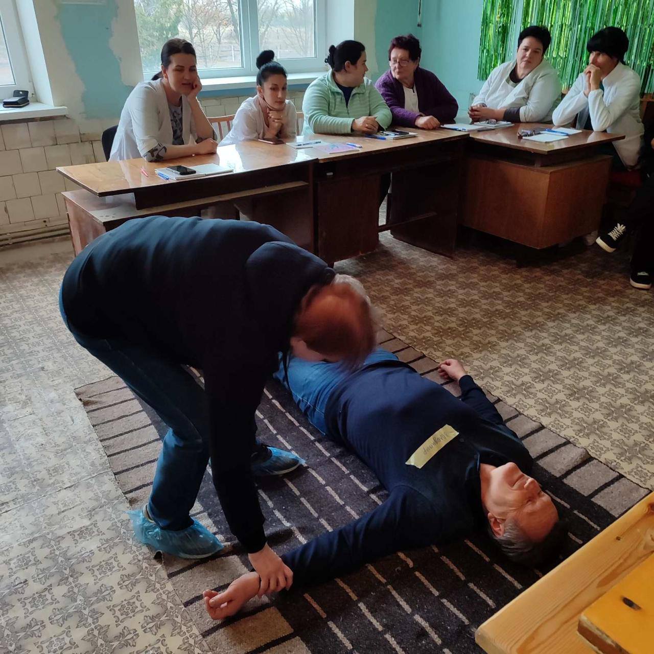 Відпрацювання навичок з надання першої домедичної допомоги. Лікарі сидять за столом, на підлозі лежить чоловік інший надає допомогу.